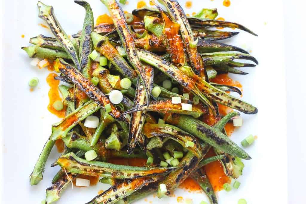 charred okra recipe topped with harissa sauce and green onions on a white plate.