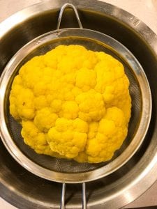 whole yellow cauliflower draining in a fine mesh strainer