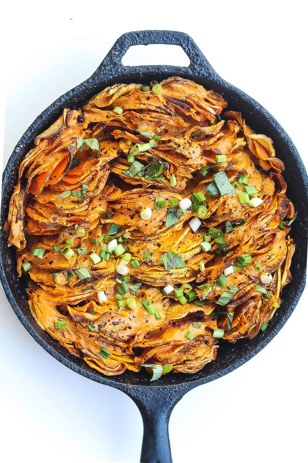 hasselback sweet potatoes in a skillet topped with green onions and red pepper sauce