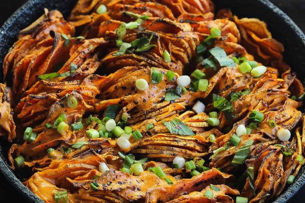 hasselback sweet potatoes in a skillet topped with green onions and red pepper sauce