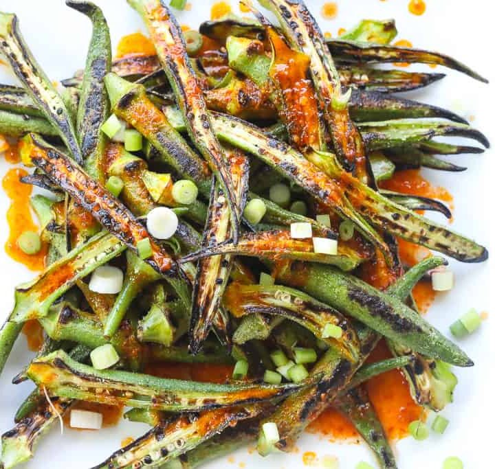 charred okra recipe topped with harissa sauce and green onions on a white plate.