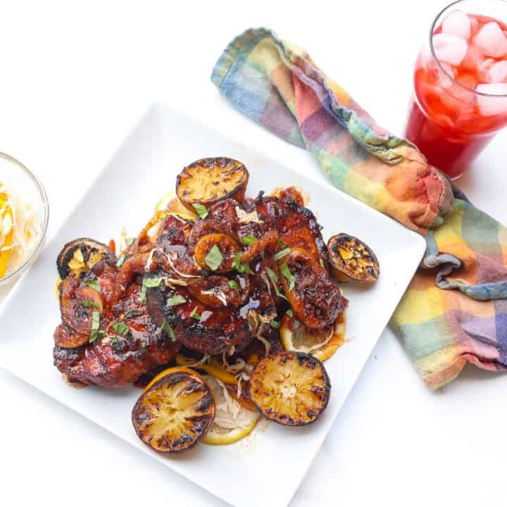 grilled pork steaks piled on white plate topped with lemons, barbecue sauce, and green onions