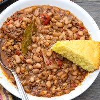 A photo of a bowl of cooked pinto beans with a garnish of a cornbread slice and small bowl of hot sauce