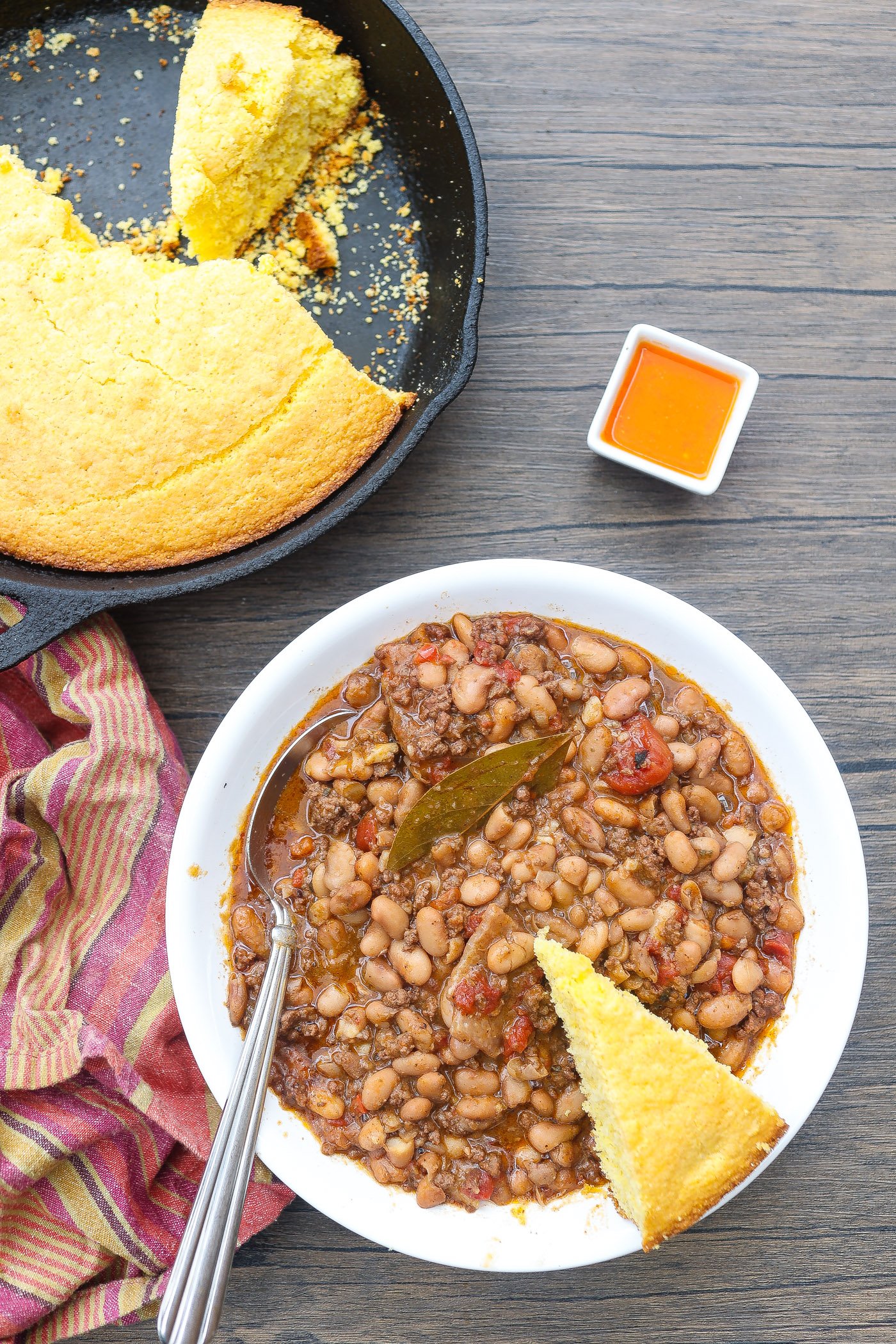 A photo of a bowl of cooked pinto beans with a garnish of a cornbread slice and small bowl of hot sauce