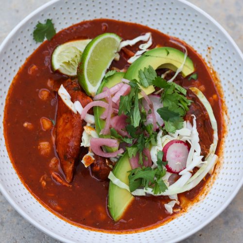 pozole rojo in a white bowl topped with avocado and pickled red onions.