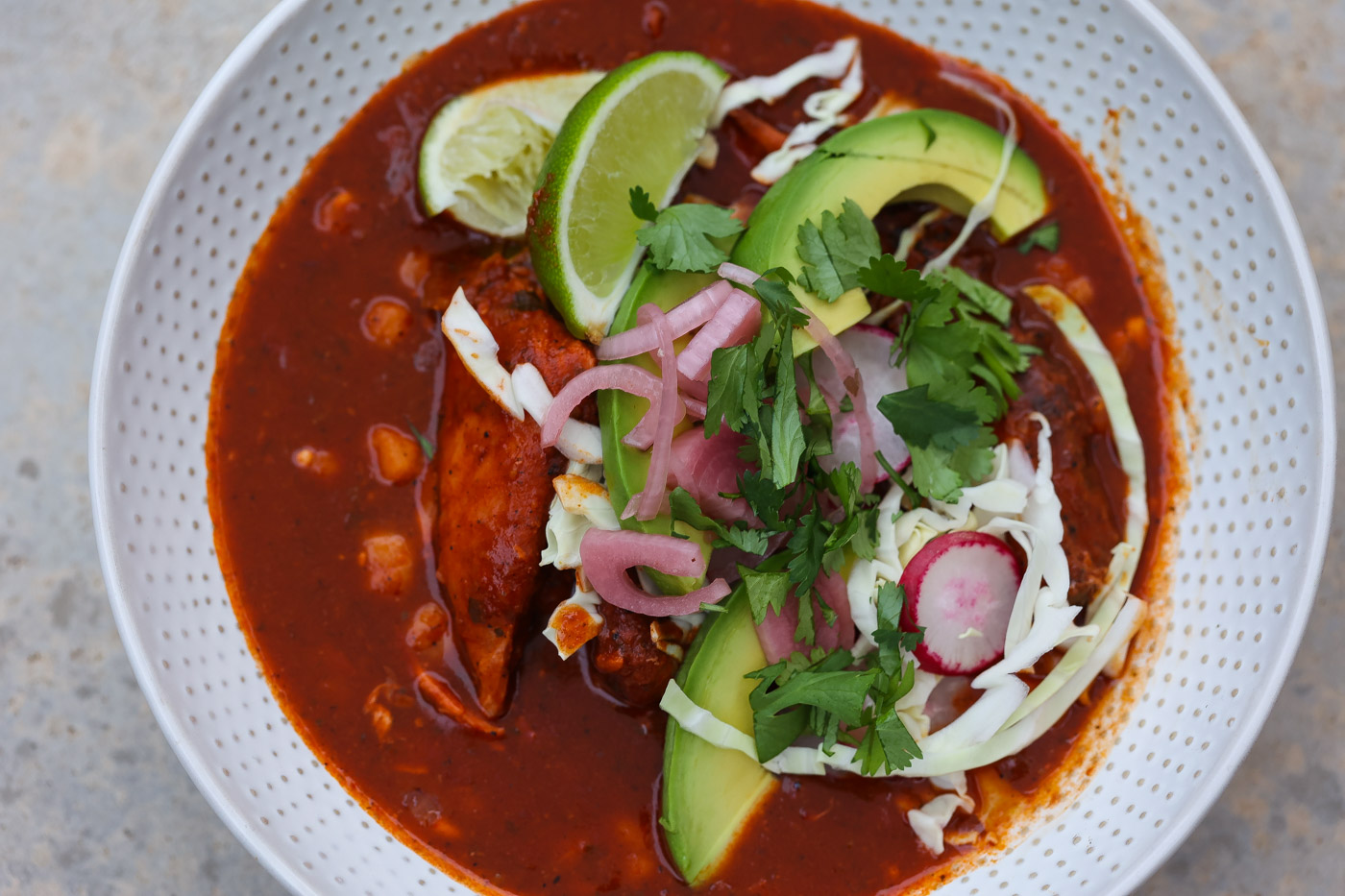 pozole rojo in a white bowl topped with avocado and pickled red onions.