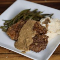 Country fried steak with brown gravy on white plate with green beans and mashed potatoes