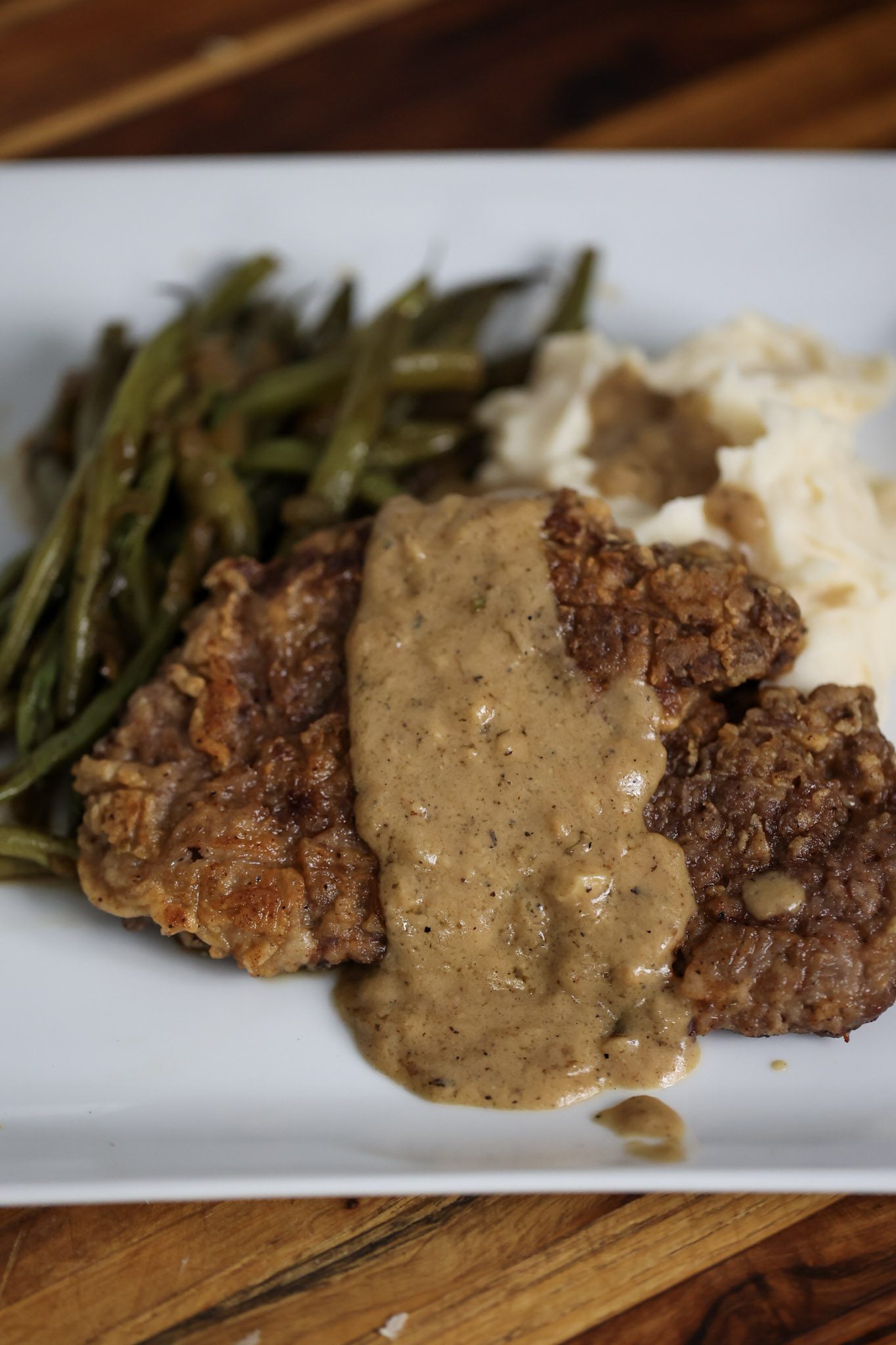 Country fried steak with brown gravy on white plate with green beans and mashed potatoes